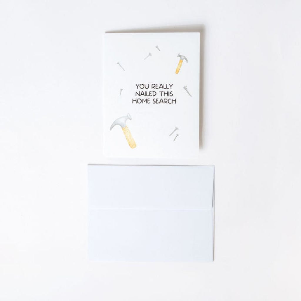 Humorous greeting card with a hammer and nail illustration and text reading "You really nailed this home search" with a white envelope on a white background