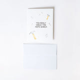 Humorous greeting card with a hammer and nail illustration and text reading "You really nailed this home search" with a white envelope on a white background