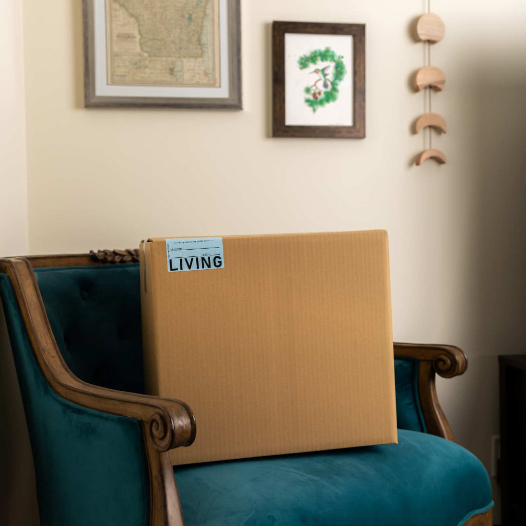 Cardboard box labeled 'LIVING' on a blue chair with framed pictures and decor on the wall.