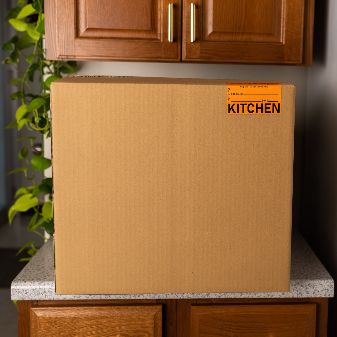 Cardboard box labeled 'KITCHEN' on a kitchen counter with cabinets and a plant in the background.