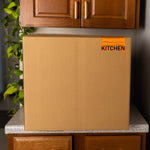 Cardboard box labeled 'KITCHEN' on a kitchen counter with cabinets and a plant in the background.