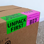 Cardboard box with 'Unpack First' and 'Bed' labels on a neutral background